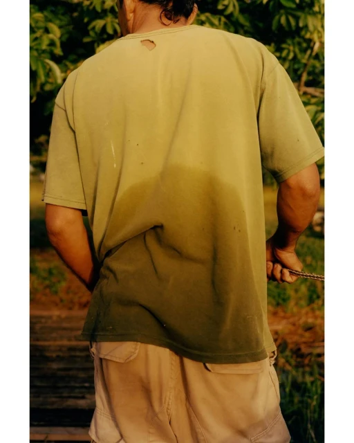 Back view of a man outdoors wearing a torn, olive t-shirt and beige cargo shorts. The t-shirt is discolored in the middle of the back.
