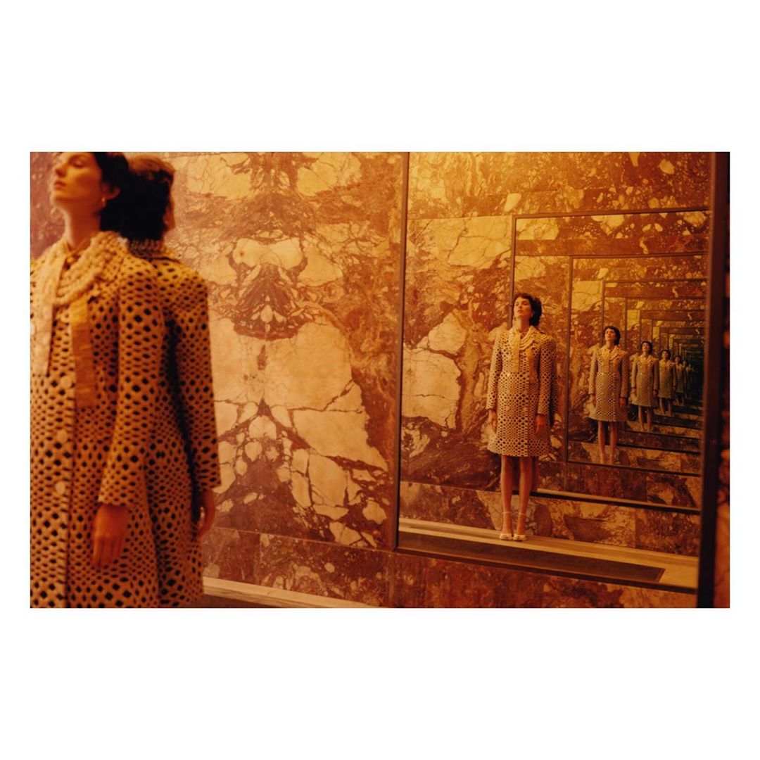 A woman in a dotted coat stands in front of mirrors, creating an infinite reflection, set against a textured, mottled wall.
