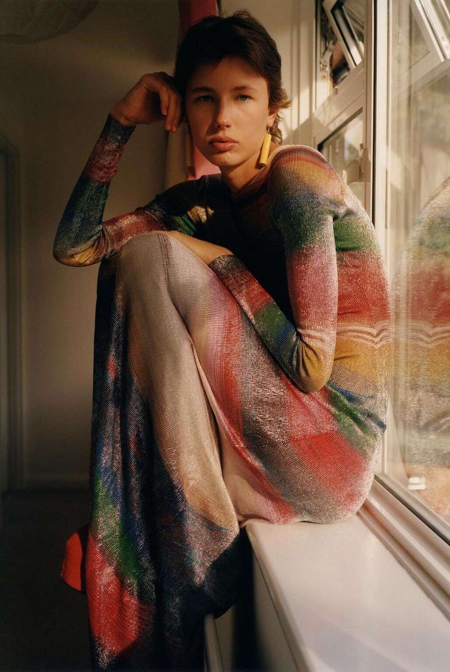 Young woman with short brown hair and rainbow dress sits near a window with natural light in an indoor environment.