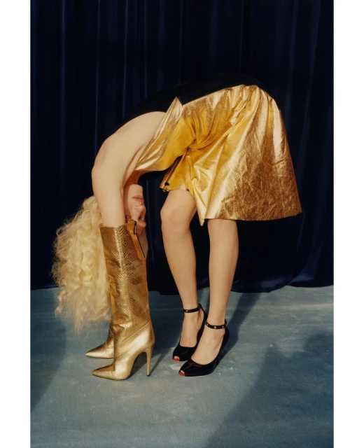 Blonde woman in gold skirt and boots bent over in studio, wearing black heels, against a dark blue curtain backdrop.