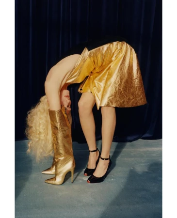 Blonde woman in gold skirt and boots bent over in studio, wearing black heels, against a dark blue curtain backdrop.