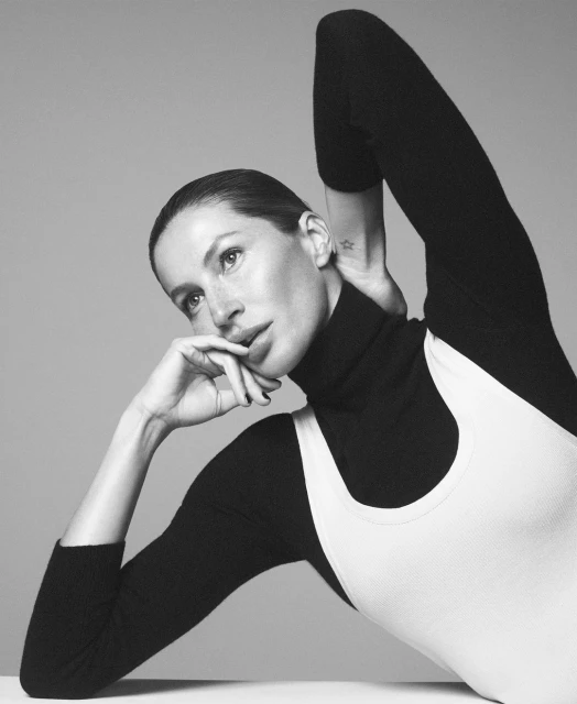 Black-and-white portrait of a woman with her hands near her face, wearing a black turtleneck and a white ribbed top, against a neutral backdrop.