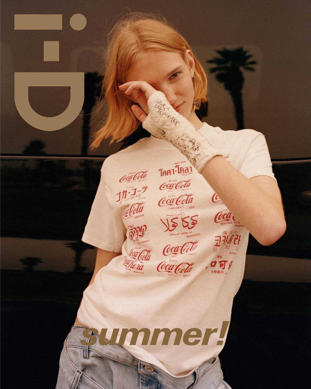 A redhead woman in a Coca-Cola t-shirt with an arm cast poses against a car with a palm tree backdrop in this fashion editorial.