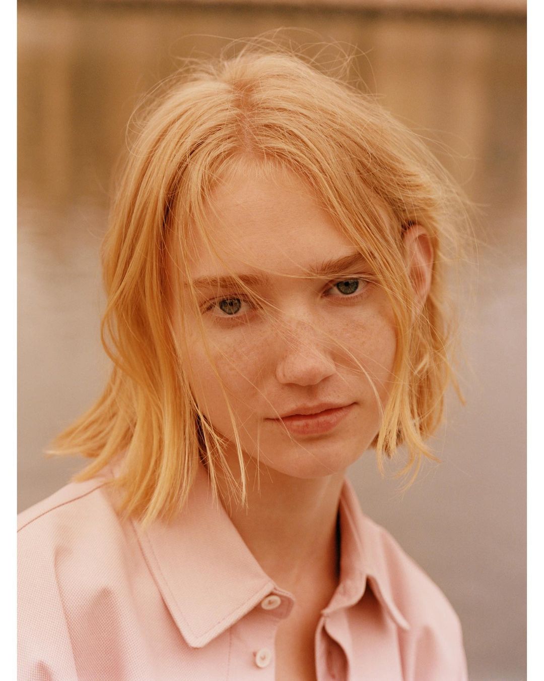 Portrait of a fair-skinned, blonde-haired woman in a pink collared shirt against a blurred outdoor background, captured in natural light.