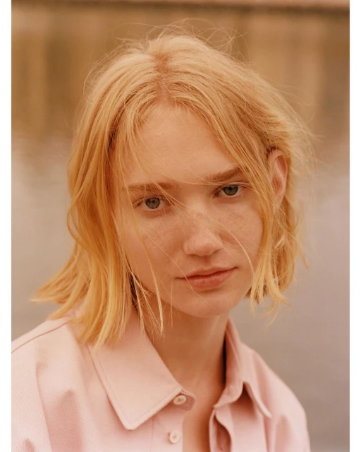 Portrait of a fair-skinned, blonde-haired woman in a pink collared shirt against a blurred outdoor background, captured in natural light.