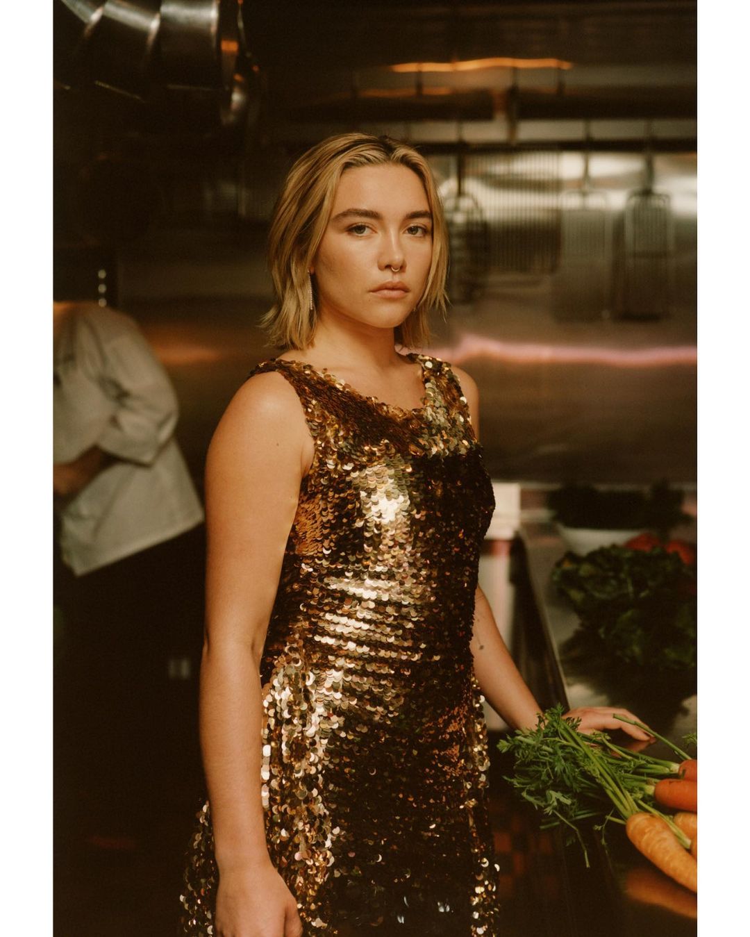 Woman in gold sequin dress holds carrots in restaurant kitchen with stainless steel. Culinary fashion and fresh food concept.