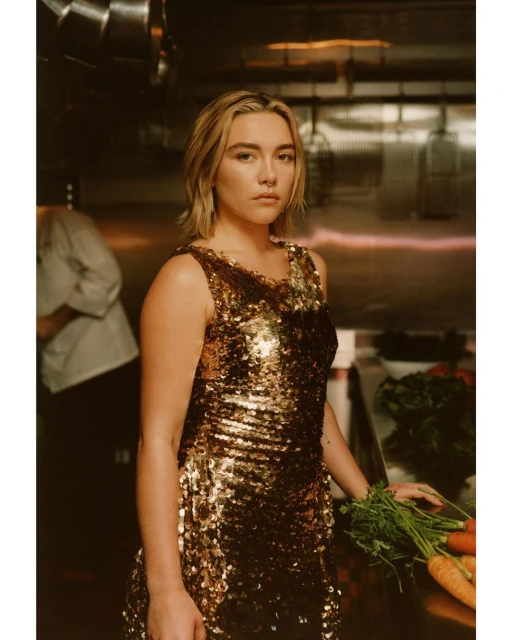 Woman in gold sequin dress holds carrots in restaurant kitchen with stainless steel. Culinary fashion and fresh food concept.