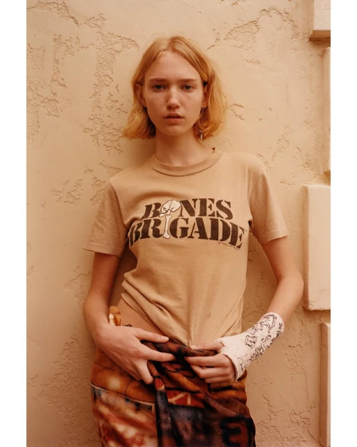 Blonde woman in Bones Brigade t-shirt poses against textured wall, wearing patterned pants. Medium shot, eye-level, soft natural light.