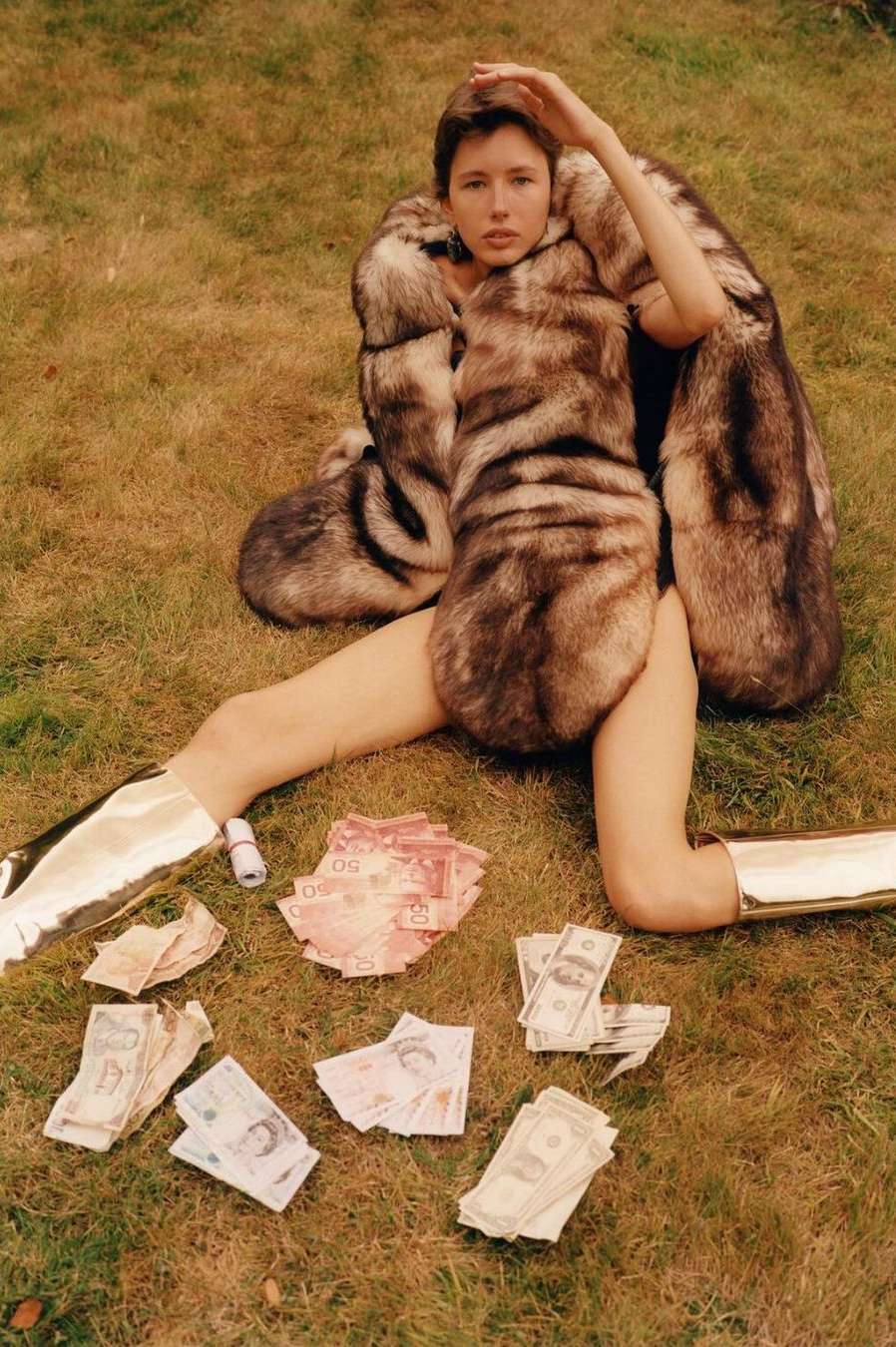 An adult woman in a fur coat and gold boots sits on the grass with scattered international paper money, creating a luxurious, playful scene.