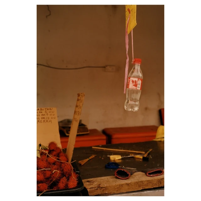 Still life shot of a market stall featuring rambutan fruit, hanging bottle, and assorted tools on a worn wooden surface in soft light.