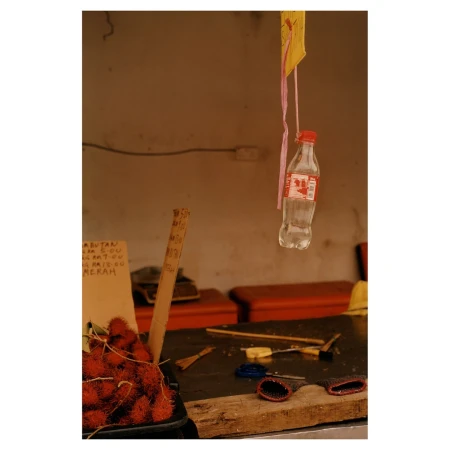 Still life shot of a market stall featuring rambutan fruit, hanging bottle, and assorted tools on a worn wooden surface in soft light.