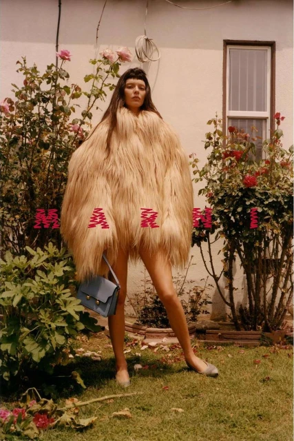 Adult woman stands outdoors in a furry, oversized coat with a handbag and shoes among rose bushes in a sunny, soft-focus fashion shot.