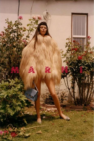 Adult woman stands outdoors in a furry, oversized coat with a handbag and shoes among rose bushes in a sunny, soft-focus fashion shot.