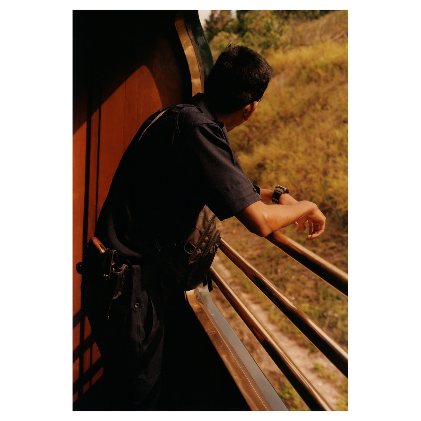 Man on train wearing dark uniform and looking out over grassy landscape. Railroad passenger with arm resting on railing.