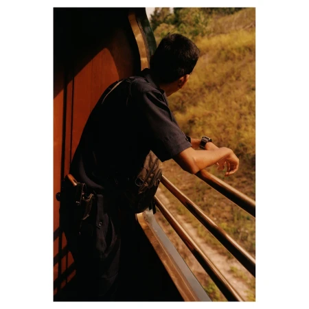 Man on train wearing dark uniform and looking out over grassy landscape. Railroad passenger with arm resting on railing.