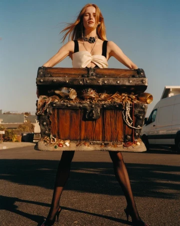 Fashionable woman poses with treasure chest in outdoor setting, wearing jewelry and fashionable dress with van and houses in background.
