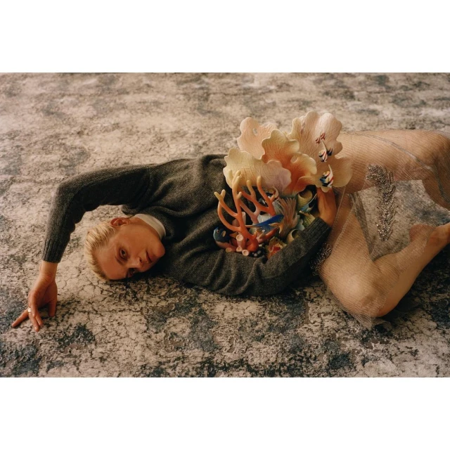Adult female with sculptural coral and flower embellishments lies on a mottled grey textile backdrop in a surreal, artistic fashion portrait.