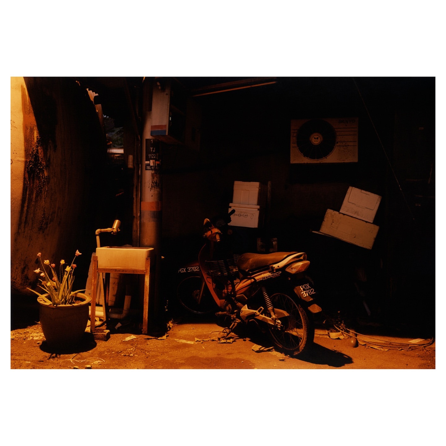 Motorbike parked outside an alleyway at night, with dim orange lighting illuminating a potted plant and white shelf.