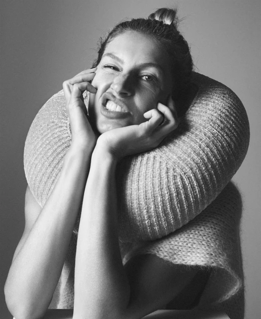 Black and white portrait of a woman with her hands near her face, wearing a large, textured sweater, and pulling a funny face.