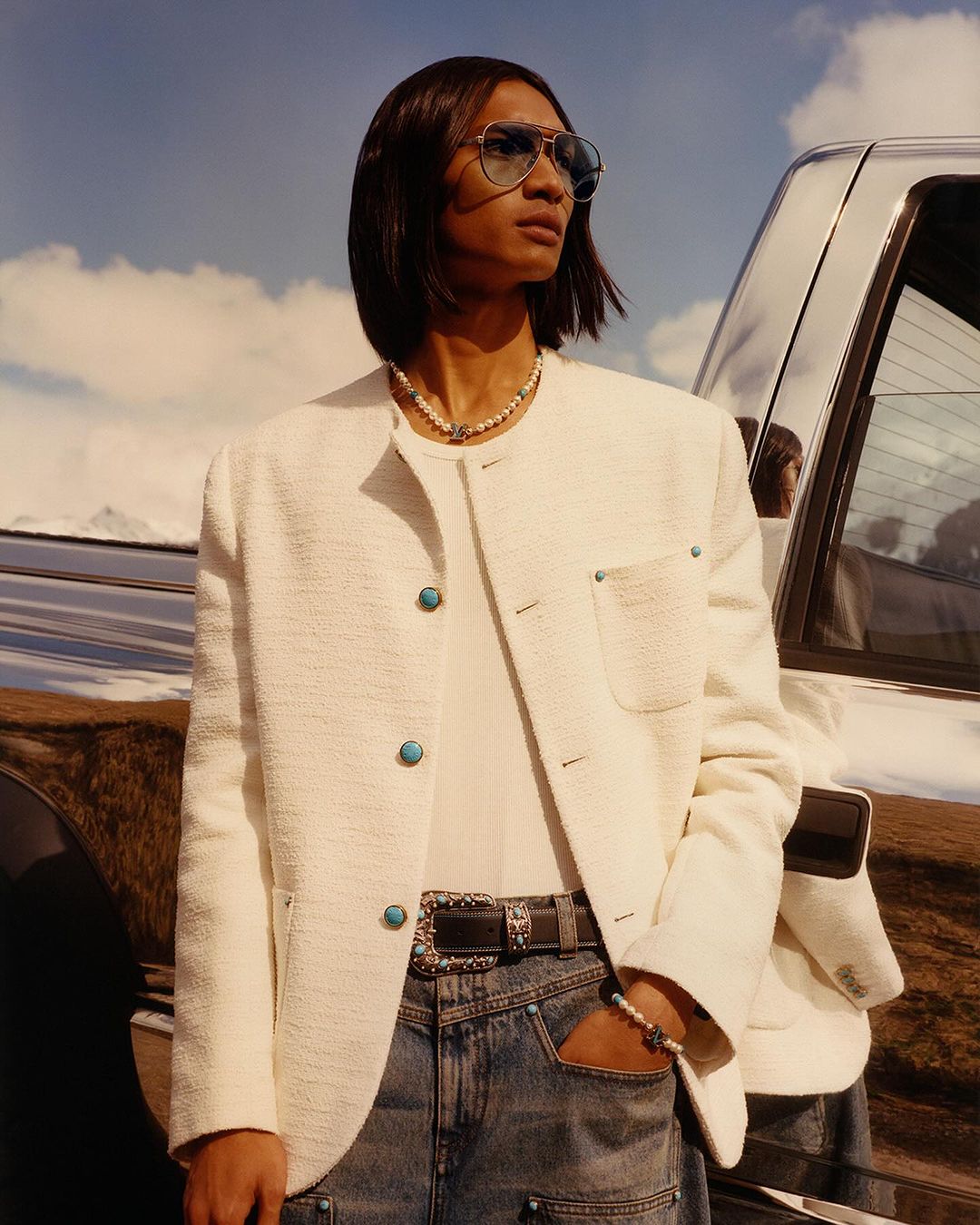 Person wearing a white jacket and blue jeans standing next to a silver truck with mountains and sky in the background, outdoors.