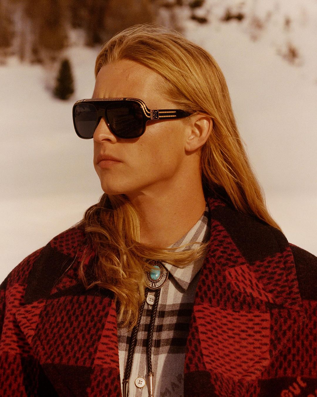 Portrait of a blond adult male wearing sunglasses and a red and black plaid coat posing outdoors against a snow-covered landscape.