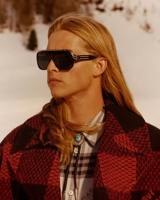 Portrait of a blond adult male wearing sunglasses and a red and black plaid coat posing outdoors against a snow-covered landscape.