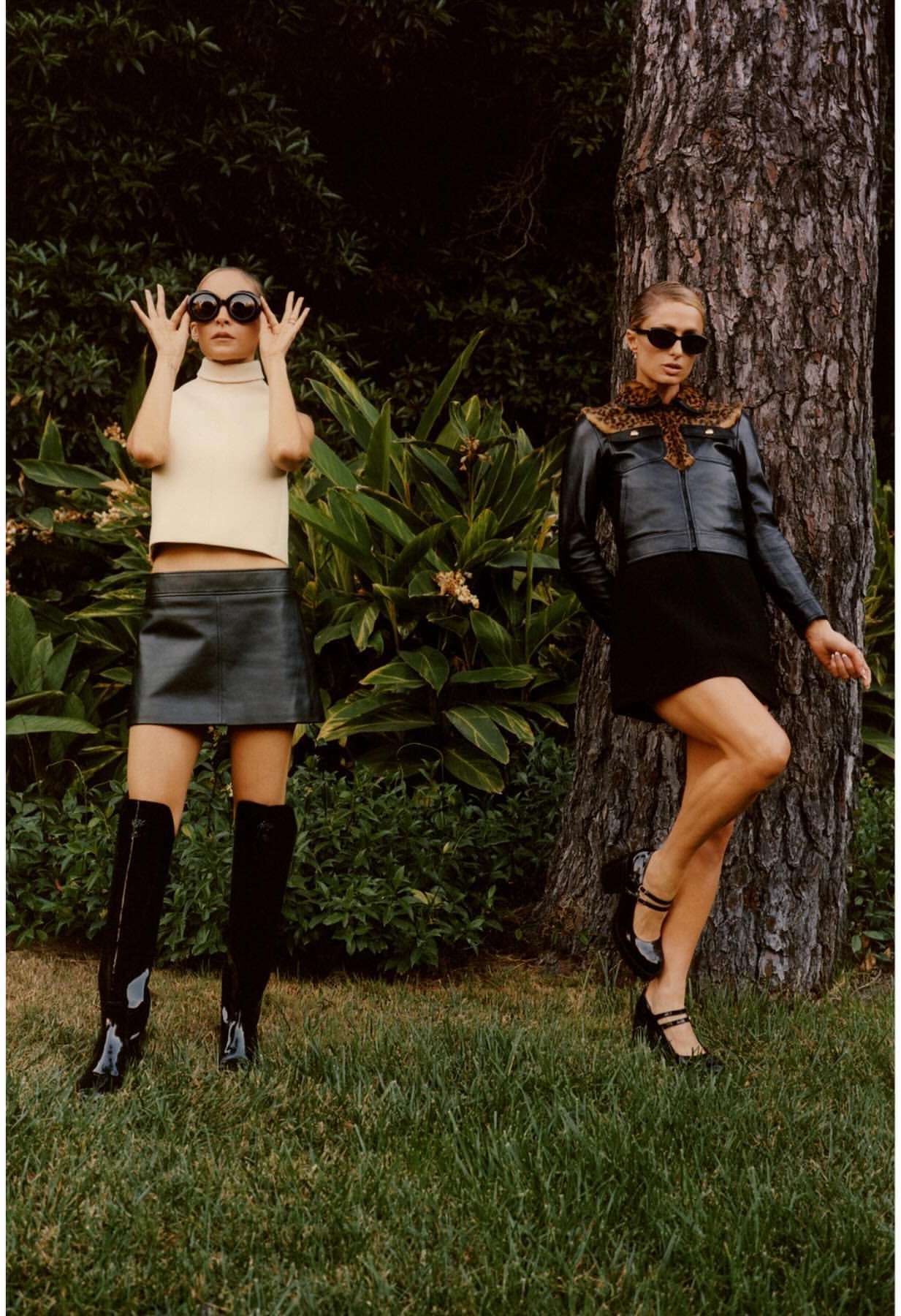 Two fashion models pose in a leafy outdoor setting. One wears a cream-colored top, black skirt, knee boots, and sunglasses; the other, a jacket, skirt, heels, and sunglasses.