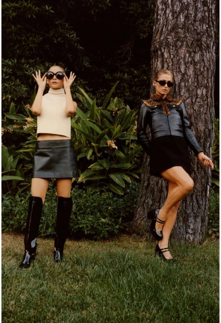 Two fashion models pose in a leafy outdoor setting. One wears a cream-colored top, black skirt, knee boots, and sunglasses; the other, a jacket, skirt, heels, and sunglasses.