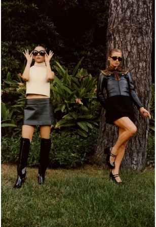 Two fashion models pose in a leafy outdoor setting. One wears a cream-colored top, black skirt, knee boots, and sunglasses; the other, a jacket, skirt, heels, and sunglasses.