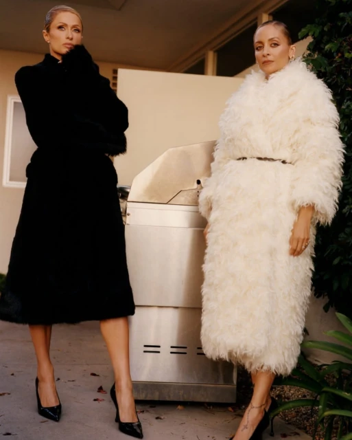Two stylish adult women posing outdoors in a fashion shot wearing fashionable fur coats and high heels, standing in front of a building.