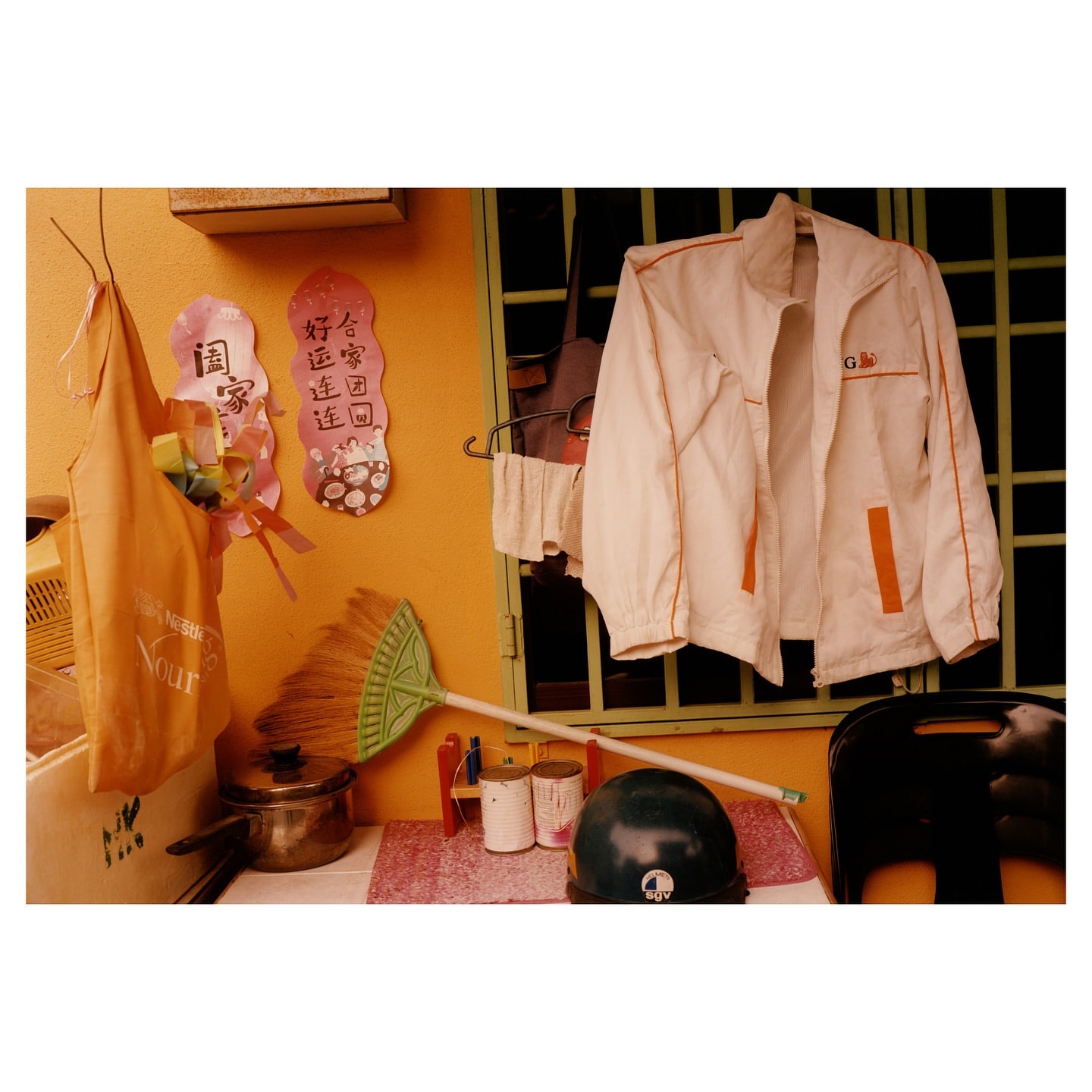 Still life featuring jacket, broom, cooking pot, helmet, and assorted household items against an orange wall with window and decorative hangings.