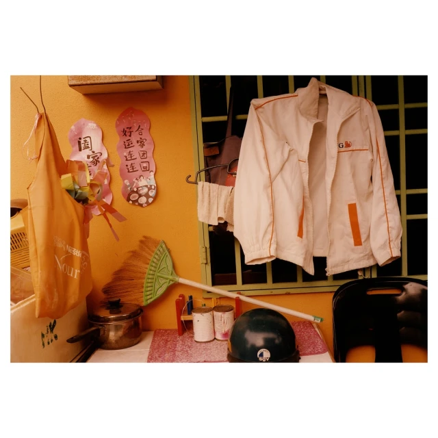 Still life featuring jacket, broom, cooking pot, helmet, and assorted household items against an orange wall with window and decorative hangings.
