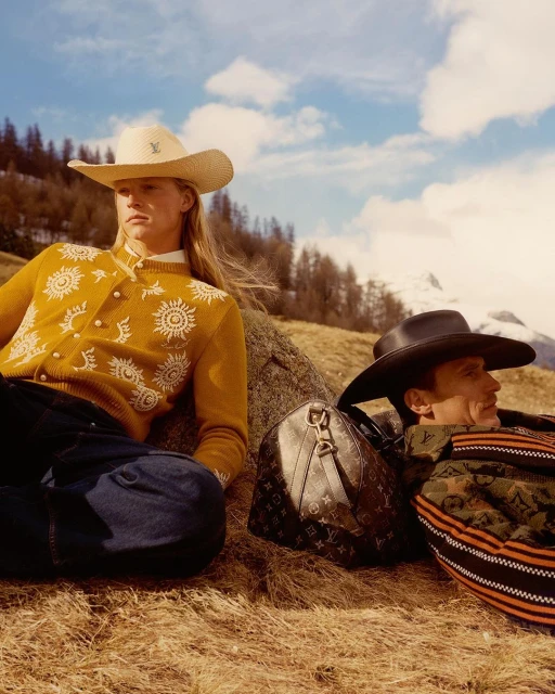 Two people wearing cowboy hats sit on a grassy hill with a Louis Vuitton duffle bag, backed by mountains and a cloudy sky.
