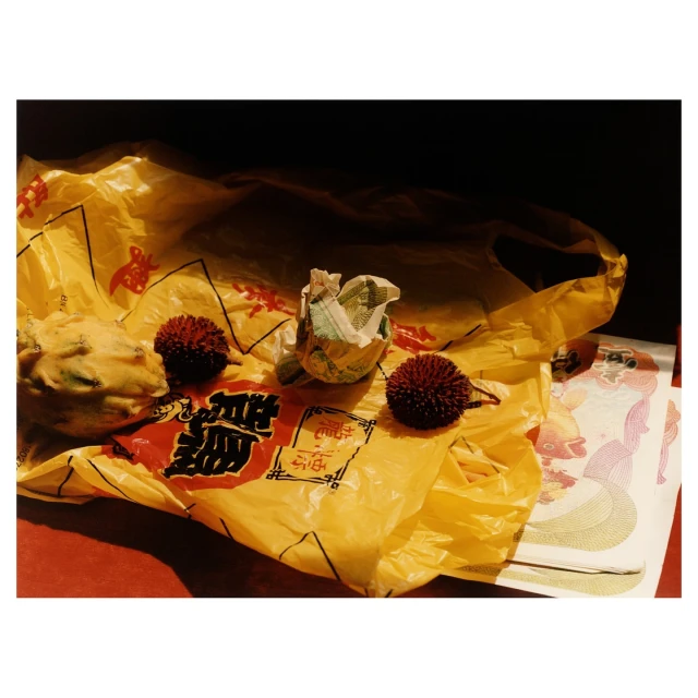 Still life shot showcasing a yellow plastic bag, several fruits including rambutans, and what appears to be joss paper on a red surface.