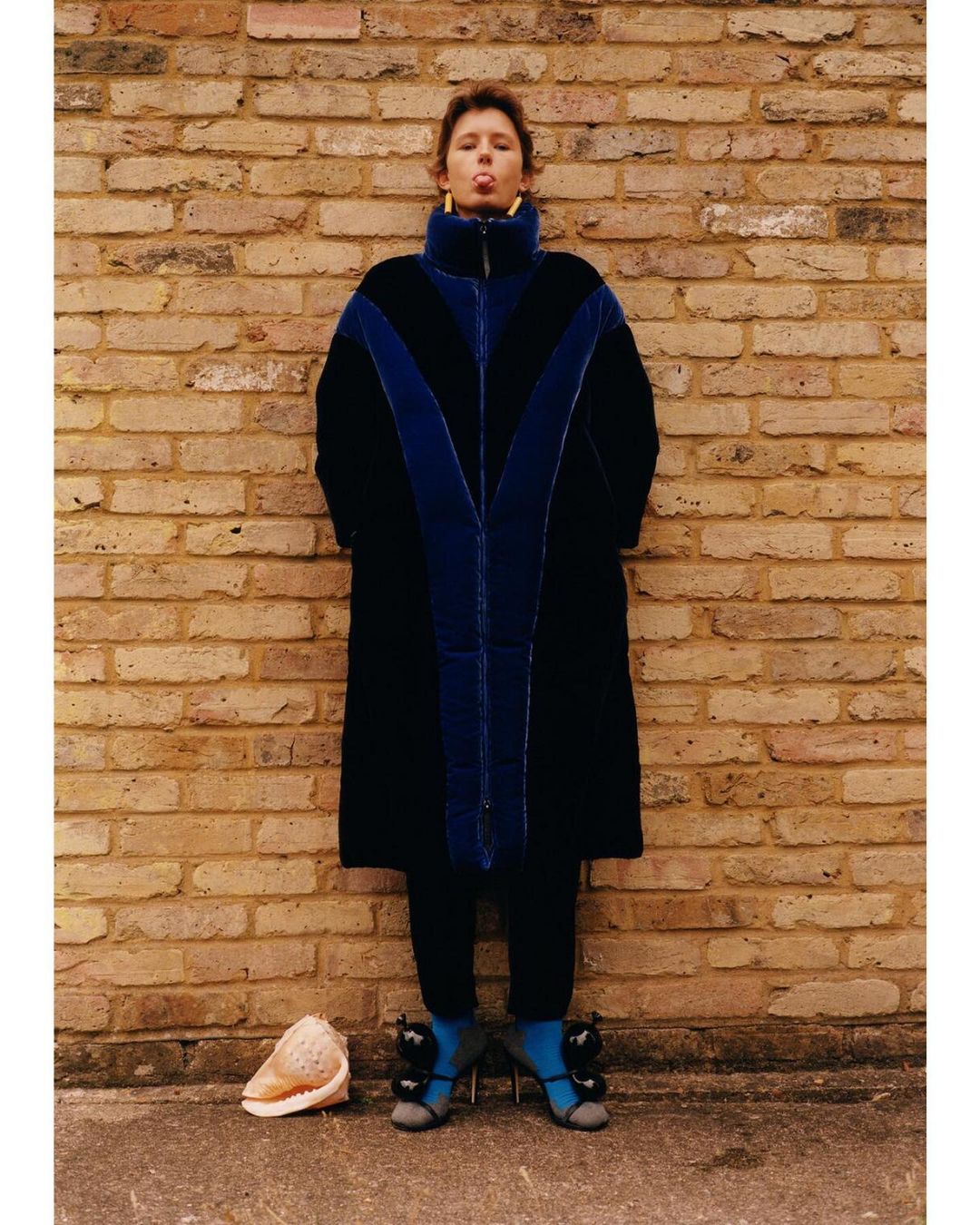 Model wearing a dark blue velvet puffer coat standing against a weathered brick wall with a large seashell on the ground.