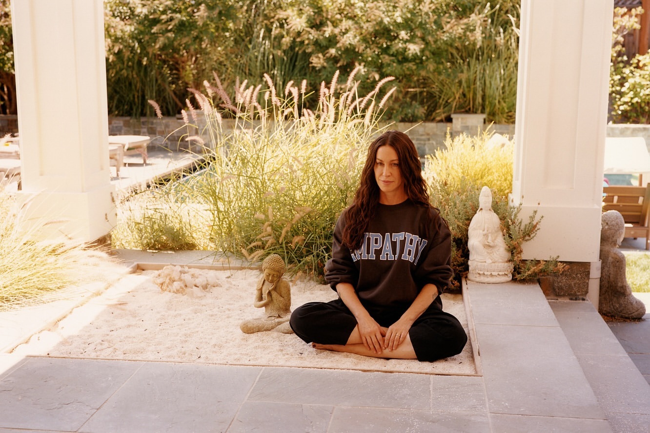 Woman sitting in a meditative pose outdoors, wearing a dark sweatshirt labeled 'EMPATHY,' on a light sand surface in a peaceful garden.