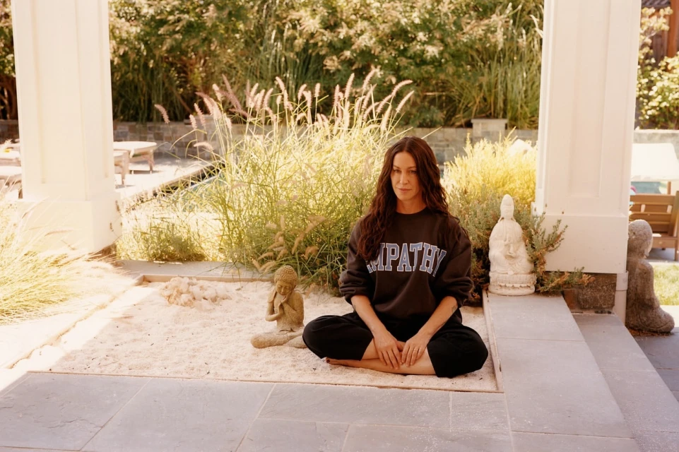 Woman sitting in a meditative pose outdoors, wearing a dark sweatshirt labeled 'EMPATHY,' on a light sand surface in a peaceful garden.