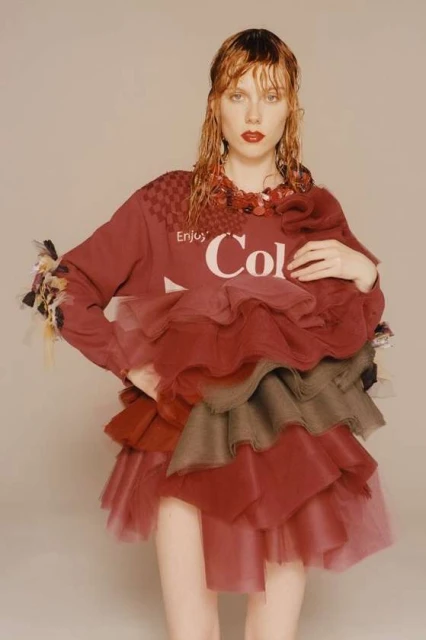 Woman posing in a burgundy sweater with the Cola-Cola logo, adorned with tiered ruffles and a checkered shoulder detail in a studio setting.