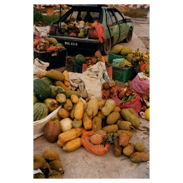 Outdoor market scene with a variety of fruits and vegetables displayed in front of a green car with its trunk open and filled with produce.