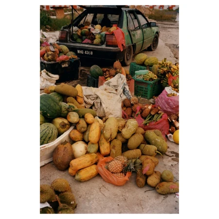 Outdoor market scene with a variety of fruits and vegetables displayed in front of a green car with its trunk open and filled with produce.