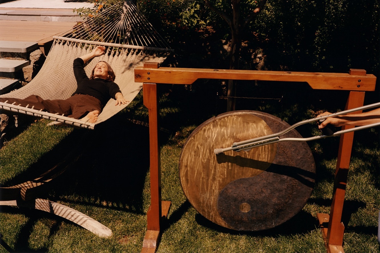 Woman rests in hammock next to gong being struck outdoors, sunny day, backyard setting, music or sound-therapy context.