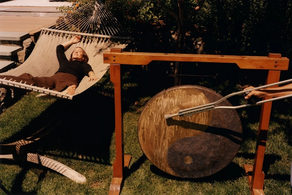 Woman rests in hammock next to gong being struck outdoors, sunny day, backyard setting, music or sound-therapy context.