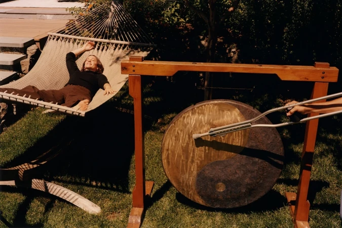 Woman rests in hammock next to gong being struck outdoors, sunny day, backyard setting, music or sound-therapy context.