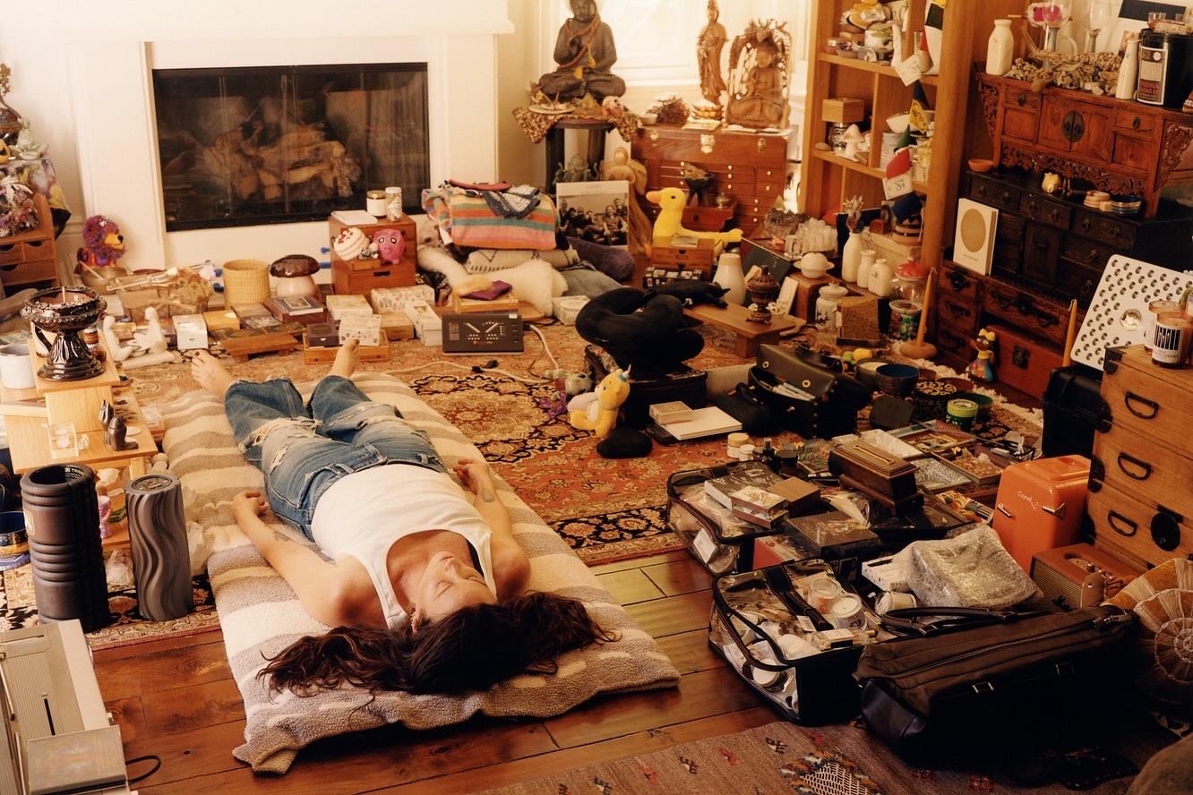 Adult woman in tank top and jeans rests on a striped mat in a room filled with assorted furniture, boxes, and objects in daylight.
