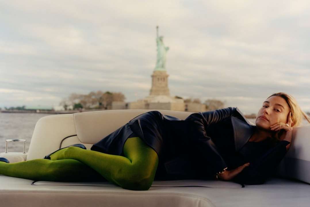 Adult woman relaxes on a yacht with the Statue of Liberty in the distance. She wears a blazer and green tights in natural light.