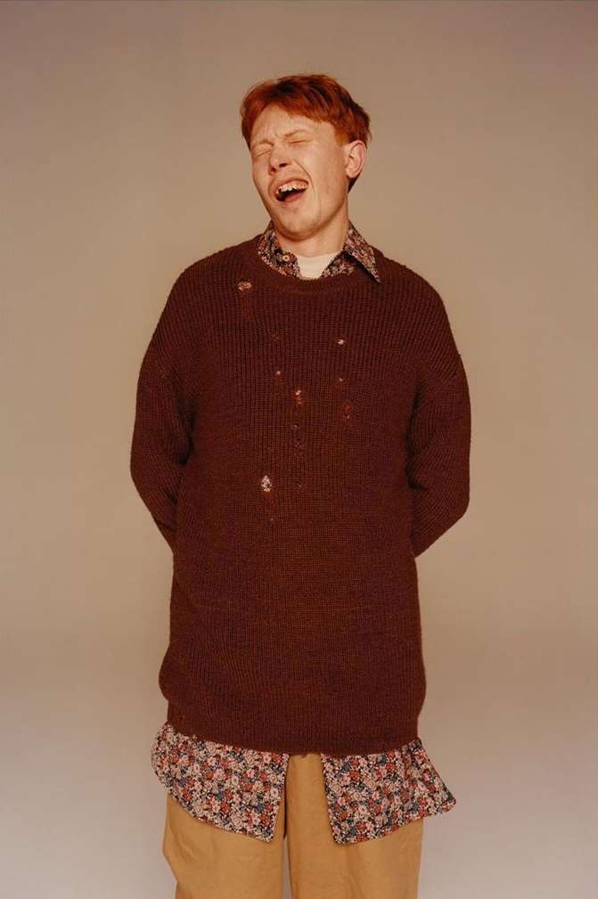 A young adult male with red hair, wearing a brown sweater and tan pants, stands in front of a neutral background with his eyes closed.
