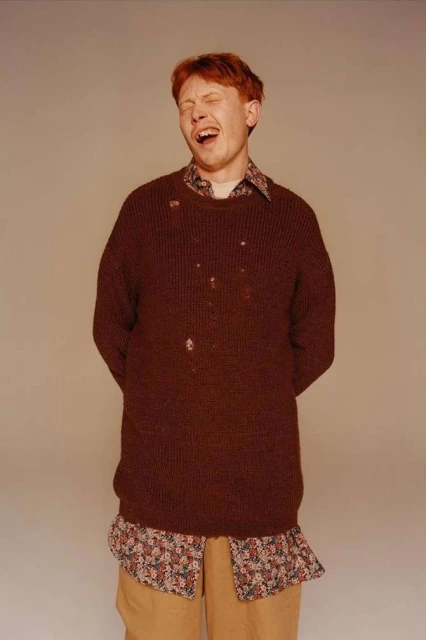 A young adult male with red hair, wearing a brown sweater and tan pants, stands in front of a neutral background with his eyes closed.