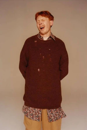 A young adult male with red hair, wearing a brown sweater and tan pants, stands in front of a neutral background with his eyes closed.