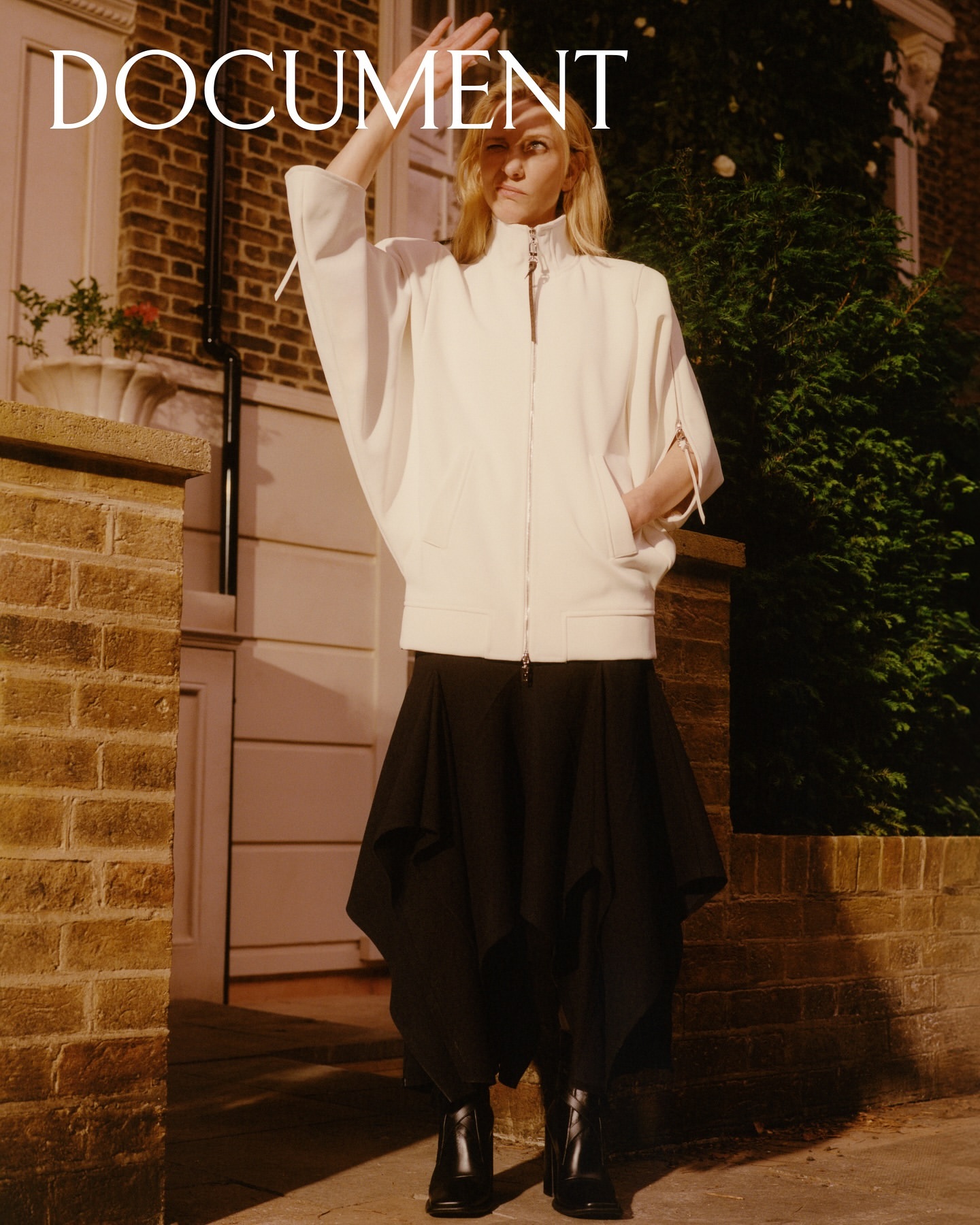 Fashion editorial shot of a blonde adult woman in a white jacket, black skirt, and black boots, posing outdoors in front of a brick building.