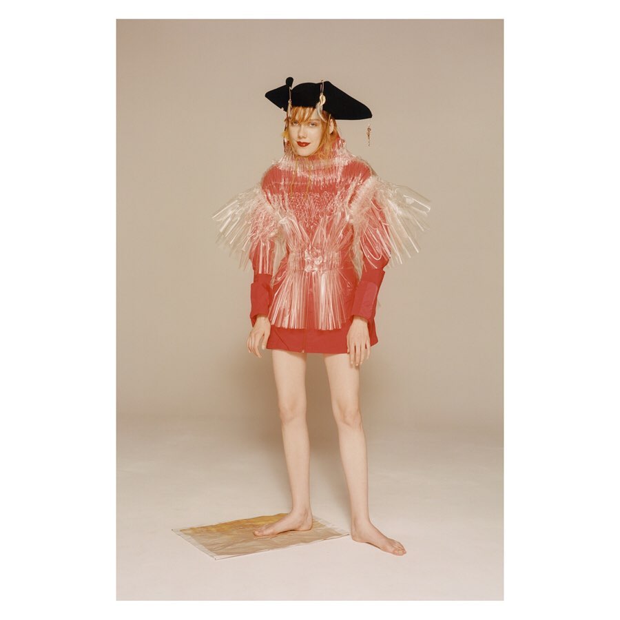 A person poses in avant-garde fashion featuring a red dress, a large brimmed hat, and a translucent vest-like piece in a studio setting.